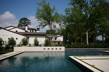 Fototapeta premium THE WOODLANDS, TEXAS - MAY 5th 2023: an outdoor pool and spa with water features on a hot sunny day. The blue water is sparkling and the Mediterranean style house is white with a terracotta roof.