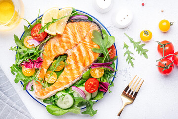 Grilled salmon steak with vegetable salad with tomatoes, arugula, onion  and lettuce on plate, white wine glass, gray table background, top view