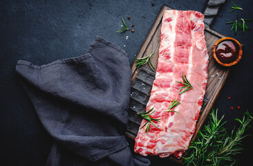 Raw pork ribs with rosemary, spices and barbecue sauce on oak wooden cutting board prepared for cooking on black kitchen table background, top view