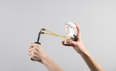 Hands holding slingshot with David bust on gray background. Modern art