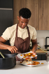 Young black man serving plantain on tomato sauce fried chicken mince and rice plate in a kitchen.