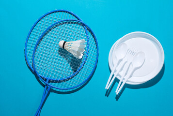 Picnic. Badminton racket with shuttlecock and plastic plate with spoons and forks on blue background with shadow