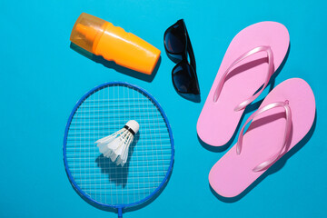 Beach vacation. Badbinton racket, shuttlecock, flip flops, sunblock and sunglasses on a blue background with shadow. Flat lay