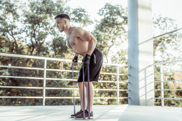 Athletic man trains muscles with fitness rubber bands outdoors. Muscular male with a naked torso exercising