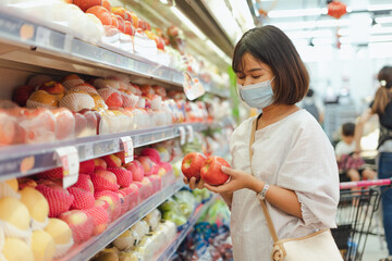 Portrait of healthy young Asian woman with casual clothes is wearing medical face mask when buy fresh organic fruit to make healthy food with new normal lifestyle  and social distancing at supermarket