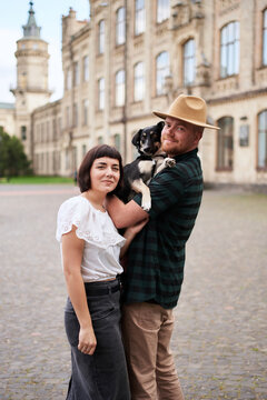 Happy couple with a shelter dog hugging in background of the old city in summer. Women with black hair and man in a hat, cute young modern couple with a rescued dog. High quality photo