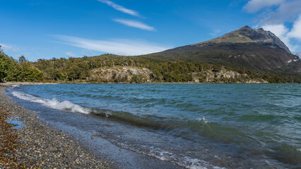 The waves of the emerald lake foam on the pebble beach. Green vegetation on the shore. A picturesque mountain on a background of blue sky and clouds. Argentina. Tierra del Fuego National Park.