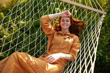 a beautiful, elegant woman lies in a long orange dress on a mesh hammock resting in nature,...