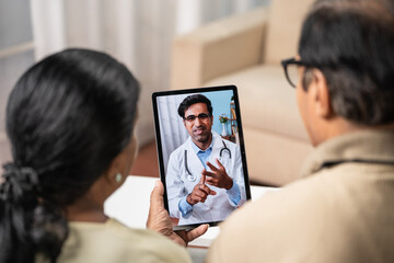 Shoulder shot of Indian Senior couple taking advice from doctor during online video call from tab at home - concept of remote health care, virtual consultation and ehealth