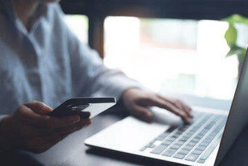 Business woman sitting at office table using mobile phone while online working on laptop computer at home office, business casual, telecommuting, working from home, close up