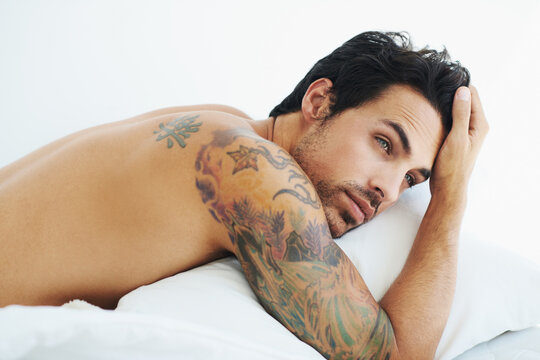 Thinking, relax and sexy with a topless man on a bed, lying in studio on a white background. Tattoo, idea and shirtless with a handsome young male model posing in a bedroom for sensuality or desire