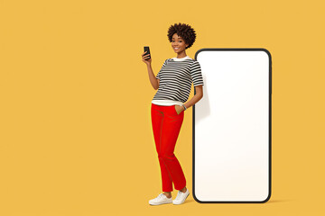 Smiling happy young afro american woman she standing near big blank screen mobile cell phone with workspace copy space mockup area use smartphone isolated on plain yellow backround.