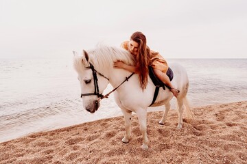 A woman in a dress stands next to a white horse on a beach, with the blue sky and sea in the background.