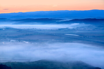 Obraz premium Aerial view of the valley in early morning mist, beautiful in the highlands. Low clouds and fog cover the sleeping meadow. Alpine mountain valley mists landscape at dawn. Serene moment in rural area