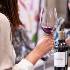 close up of female hand supporting a wineglass with red wine. Friends and wine bottles on the background. Wine tasting with friends