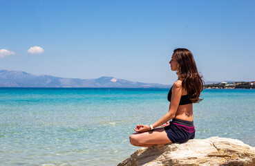 Fototapeta na wymiar Young beautiful slim woman with long brown hair is meditating in yoga position on a rock by the Aegean sea in a sunny day. Healthy lifestyle concept