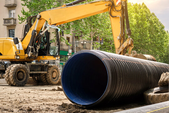 Working excavator on the roadway street. Close-up of large plastic corrugated pipe for water supply against the background of construction equipment.