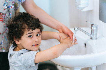 Handsome caucasian curly little boy washing hands at medical office looks back at camera smiles. Cropped image of nurse helping kid to clean hands before medical exam. Health and medicine.