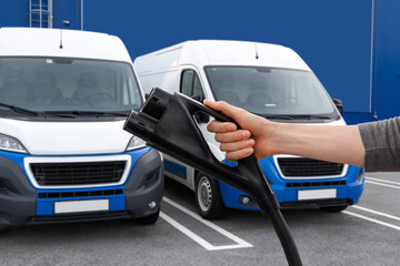 Hand with electric vehicles charging plug on a background of electric delivery vans 