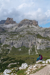 Fototapeta premium A girl, a tourist in blue clothes with a crimson backpack, sits with her back on a stone and looks at the majestic Dolomite mountains in Italy. Mountain View