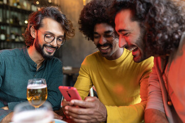 Three diverse young adult friends using mobile phone sitting on table at bar. Technology and male friendship concept.