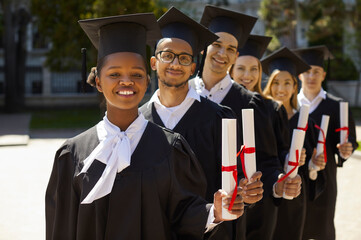 Outdoor portrait of happy diverse university students on graduation day. Group of joyful proud mixed race multiethnic graduates in black caps and gowns standing in row, holding diplomas and smiling
