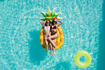 Top view of beautiful girls in Santa's hats relaxing on inflatable pineapple mattress in pool....