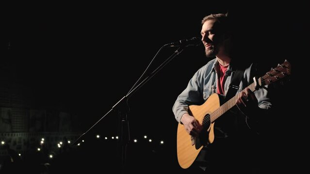 A musician is playing guitar on a large stage with audience members holding flashlights in the background. The auditorium is filled with beautiful flashing lights.