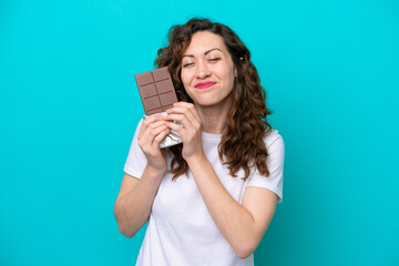 Young caucasian woman isolated on blue background taking a chocolate tablet and happy