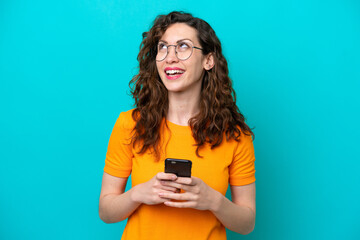 Young caucasian woman isolated on blue background using mobile phone and looking up