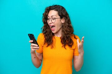Young caucasian woman isolated on blue background looking at the camera while using the mobile with surprised expression