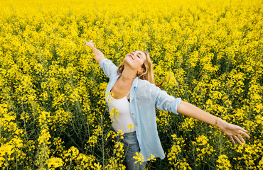 Fototapeta premium Beautiful young blonde cheerful woman, with outstretched arms, amidst a field of blooming yellow rapeseed flowers