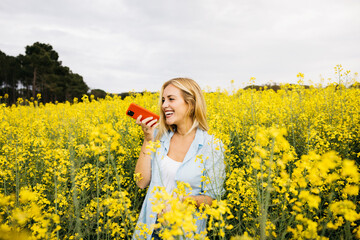 Fototapeta premium Beautiful young blonde cheerful woman, sending a voice message using a red phone, amidst a field of blooming yellow rapeseed flowers