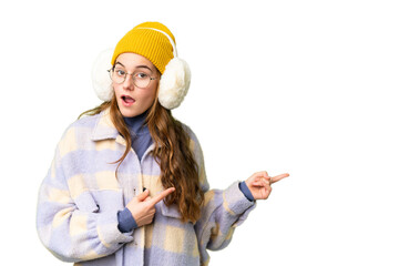 Teenager girl wearing winter muffs over isolated chroma key background surprised and pointing side