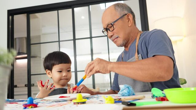 Cheerful Asian Japanese Senior man and little child boy in apron Enjoying painting art at home together. Happy Grandfather coloring on Grandchild hands. Positive emotion. Laughing