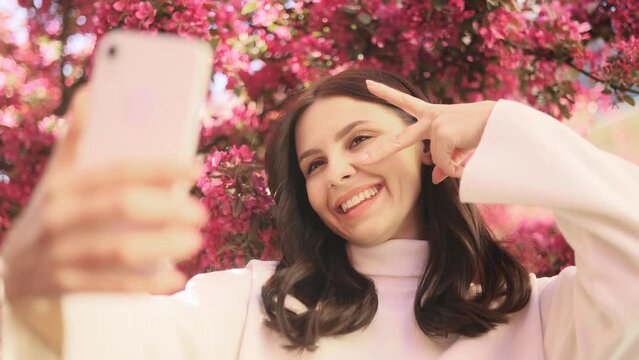 Portrait of smiling dark haired woman posing takes selfies and streams video to social media from her smartphone with pink flowers blossom on the background outdoors