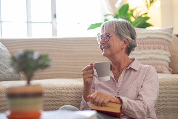 Attractive senior woman sitting on the floor at home relaxing with a coffee cup and a book