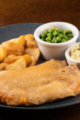 plate of fish and chips with sliced ​​fried fish and pub table closeup with tartar sauce and green peas closeup portrait on breading