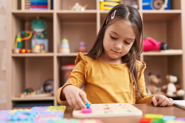 Adorable hispanic girl playing with maths puzzle game sitting on table at kindergarten