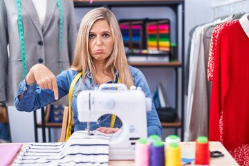 Blonde woman dressmaker designer using sew machine pointing down looking sad and upset, indicating direction with fingers, unhappy and depressed.