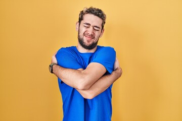 Hispanic man with beard standing over yellow background hugging oneself happy and positive, smiling...
