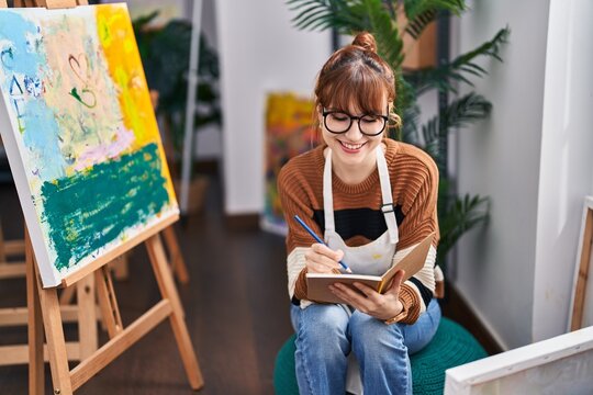 Young woman artist smiling confident writing on book at art studio