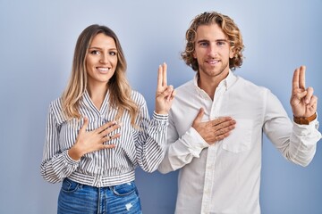Young couple standing over blue background smiling swearing with hand on chest and fingers up,...