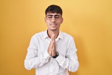 Young hispanic man standing over yellow background begging and praying with hands together with hope expression on face very emotional and worried. begging.