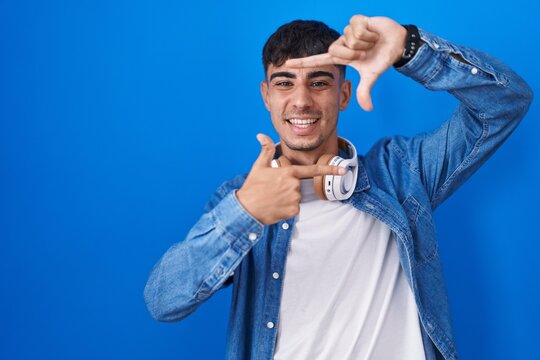 Young hispanic man standing over blue background smiling making frame with hands and fingers with happy face. creativity and photography concept.