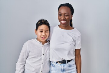Young mother and son standing together over white background winking looking at the camera with...