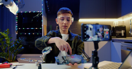 Smart teenage boy vlogger look at camera phone and recording podcast. Male guy holding electronic board and showing to followers at home. Education and modern technologies concept.
