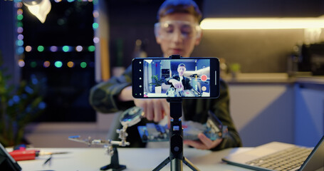 Focus on screen of smartphone. Young man is live streaming a training blog about computer motherboard circuit talking and holding device in eveting at home.
