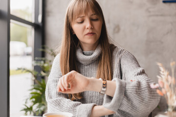 Fototapeta Checking time. Serious young worried woman sitting in a cafe and looking at her watch while waiting for her friend with a smart phone being placed on the table by her side. obraz