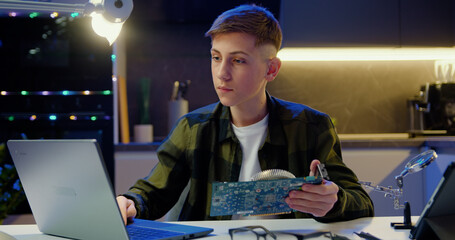 Attractive Modern teen boy in protective glasses considering computer board in his hands and...